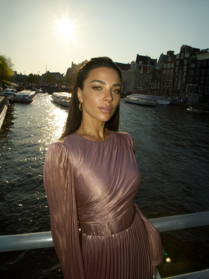 Woman in a dusty rose pleated dress standing by a canal with boats and buildings in the background.