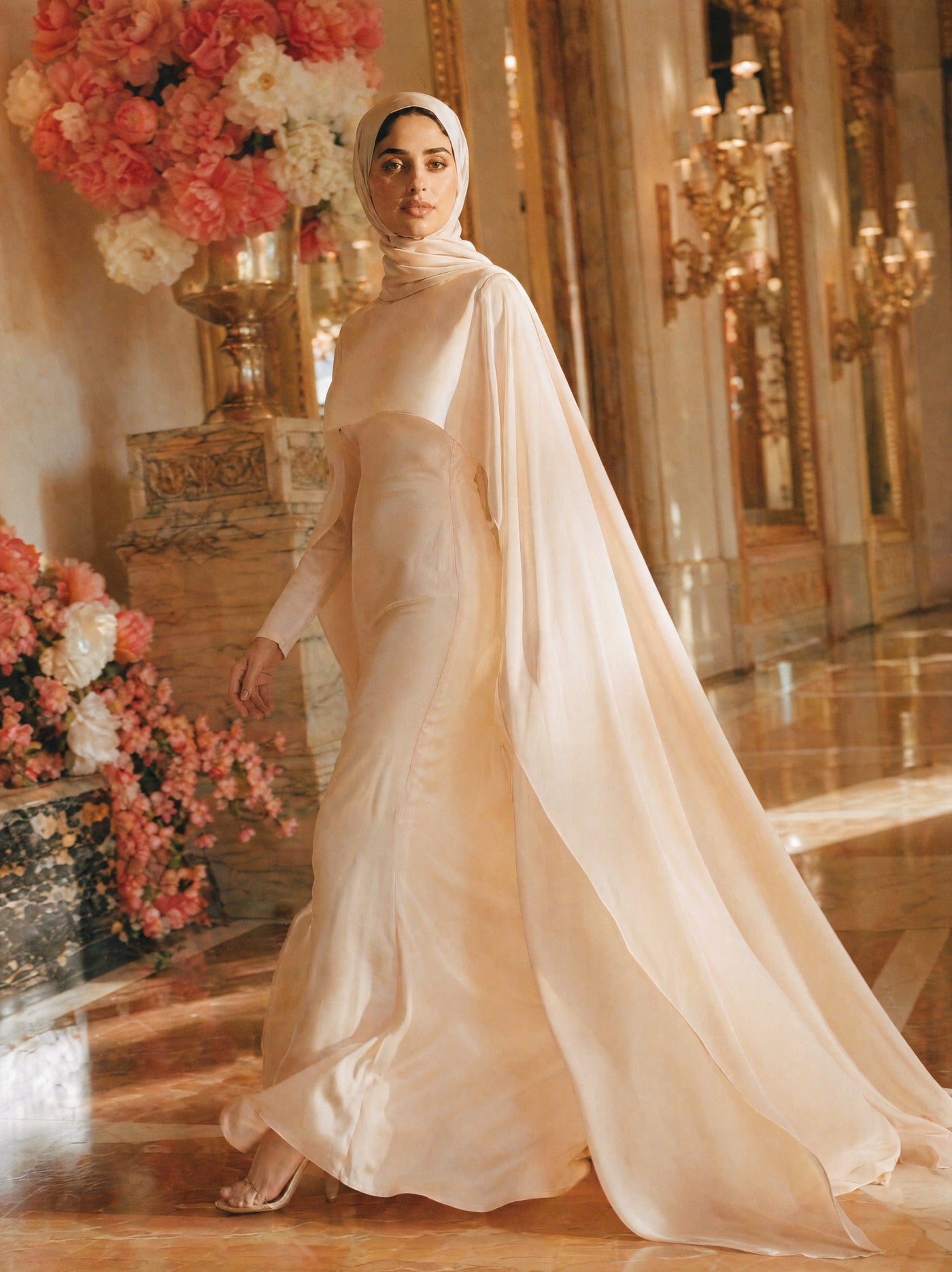 Mystic Modesty SS26— Yasmine Beige chiffon cape gown, sunlit palatial corridor mid-stride with rose-gold sconces, modest evening wear, Vogue editorial film aesthetic